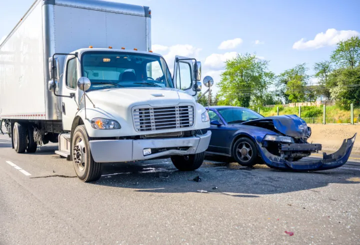 A white semi-truck and a black car are involved in a collision on a road, with visible front-end damage to the car and bumper debris on the pavement.