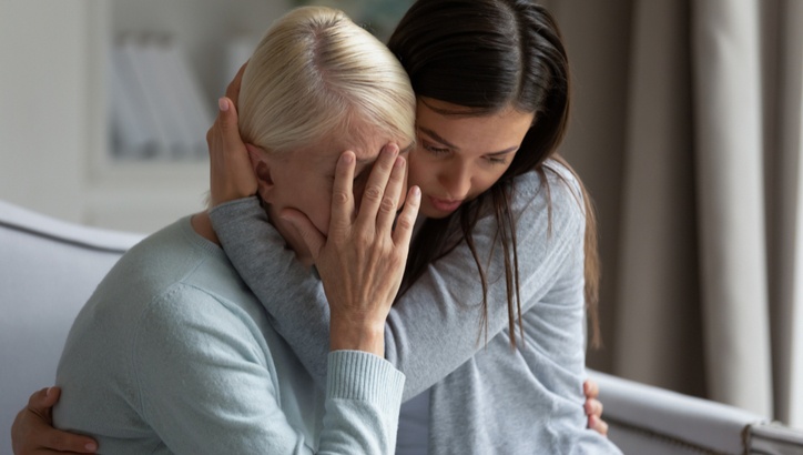 A young woman hugs and comforts an older woman who is covering her face with her hand, sitting together on a sofa indoors.