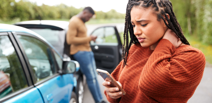 Woman holding her neck and looking at her phone, standing next to a blue car after an accident, with another person using a phone near a second vehicle in the background.