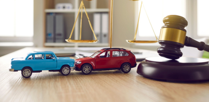 Two toy cars, one blue and one red, depict a collision in front of a judge’s gavel and a set of scales, symbolizing legal proceedings related to a car accident.