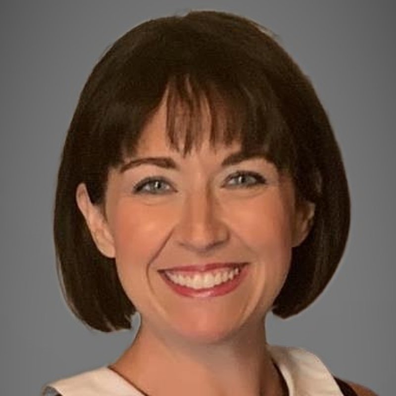 A woman with short brown hair and bangs smiling, wearing a white blouse, against a plain gray background.