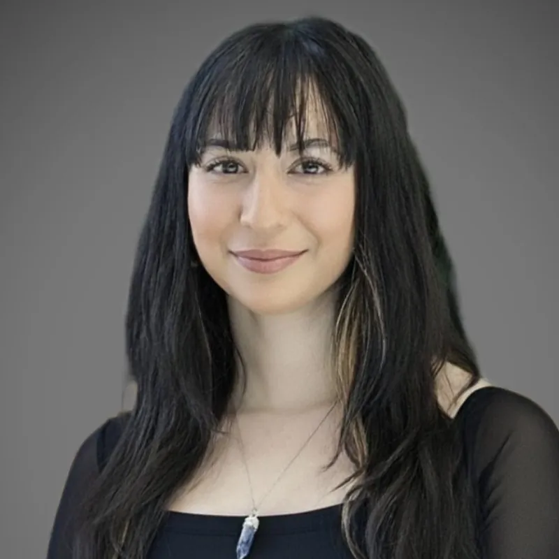A woman with long black hair and bangs, wearing a black top and a pendant necklace, poses in front of a plain grey background.