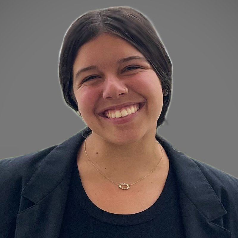 A person with straight, dark hair, wearing a black top and jacket, smiles at the camera against a plain gray background.