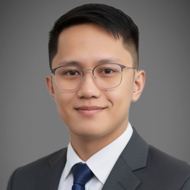 A young man wearing glasses, a dark suit, white shirt, and navy tie poses in front of a plain gray background.