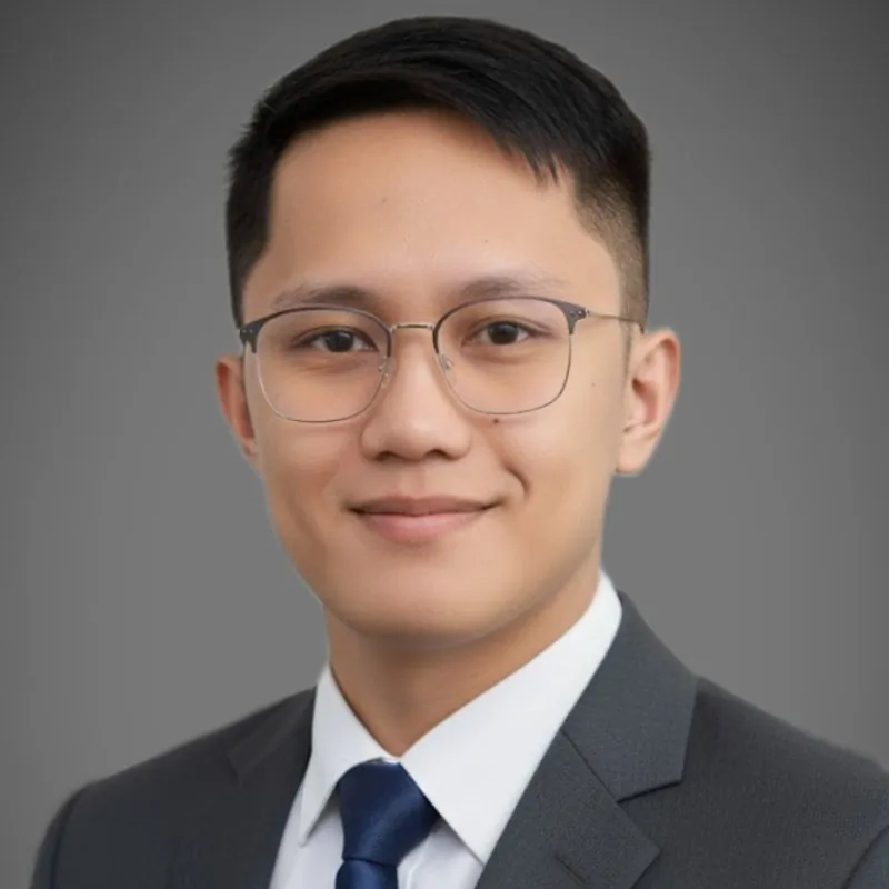 A young man wearing glasses, a dark suit, white shirt, and navy tie poses in front of a plain gray background.