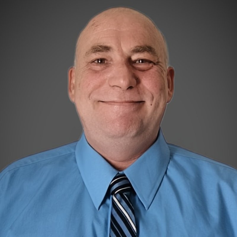 A middle-aged man with a bald head smiles at the camera, wearing a light blue dress shirt and a striped tie, against a plain gray background.