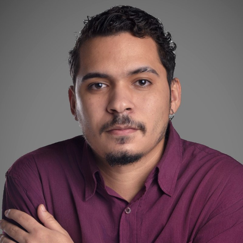 A man with short curly hair, a goatee, and a maroon button-up shirt looks directly at the camera against a plain gray background.
