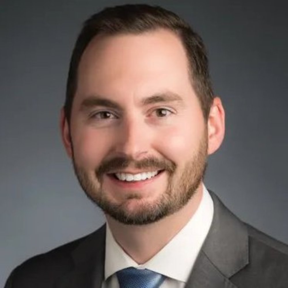 A man with short brown hair, a beard, and mustache is wearing a gray suit, white shirt, and blue tie, smiling in front of a dark, plain background.