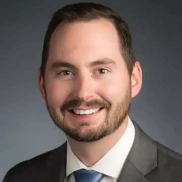 A man with short brown hair, a beard, and mustache is wearing a gray suit, white shirt, and blue tie, smiling in front of a dark, plain background.