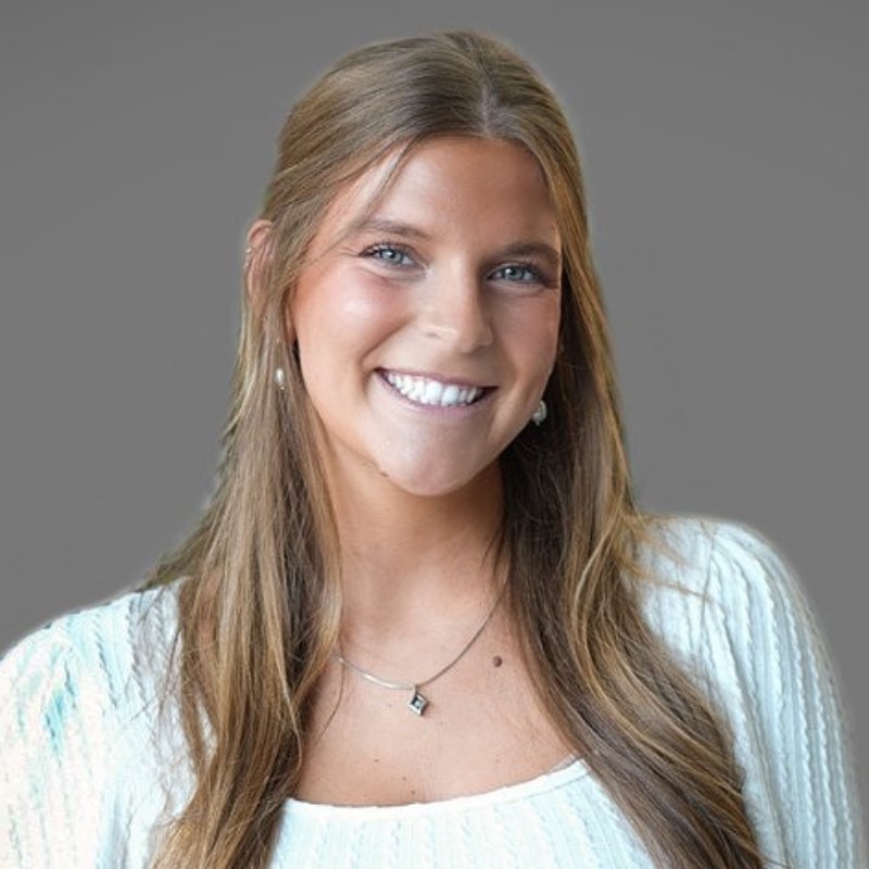 A young woman with long light brown hair wearing a white top, small hoop earrings, and a necklace, smiling in front of a plain gray background.