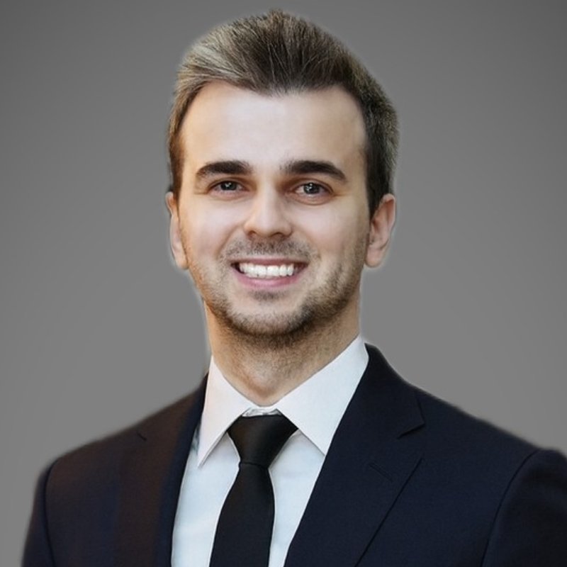 A man in a dark suit, white shirt, and black tie smiles at the camera against a plain gray background.