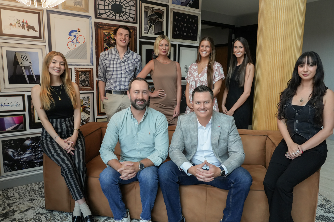 Eight people are posing indoors in front of a gallery wall; three are seated on a brown couch, and five are standing behind them, all dressed in business casual attire.