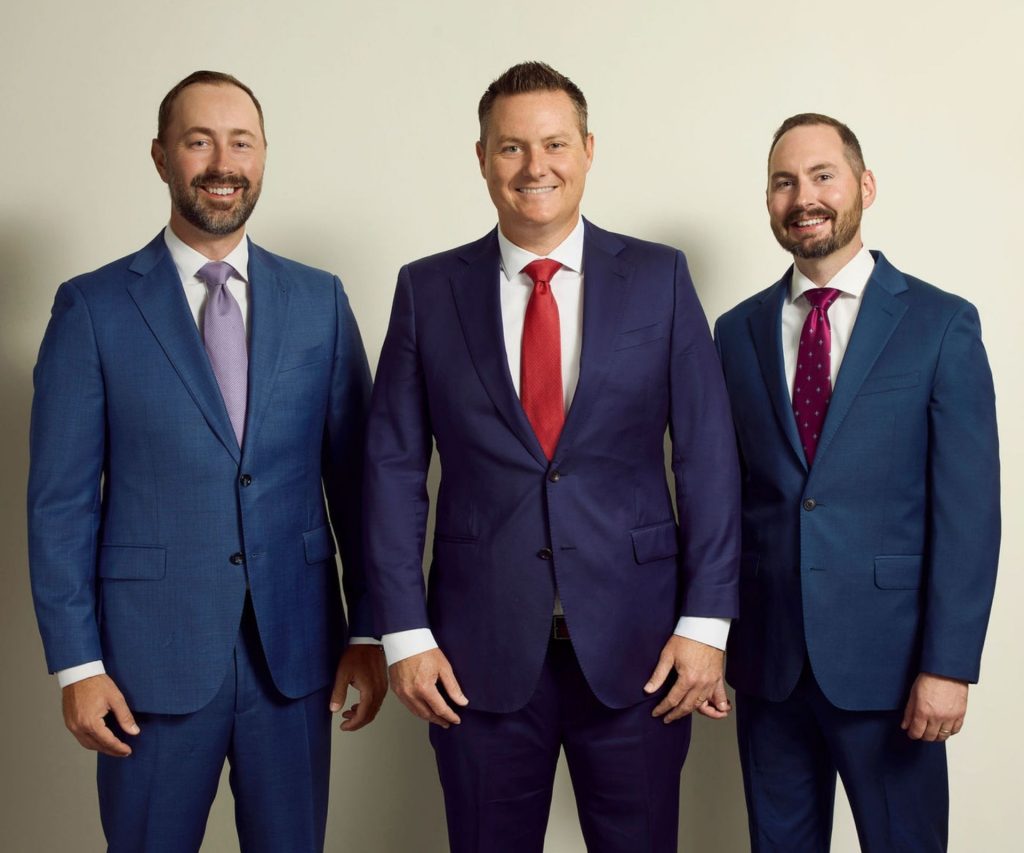 Three men in blue suits stand side by side against a plain light-colored background, all smiling and wearing ties.