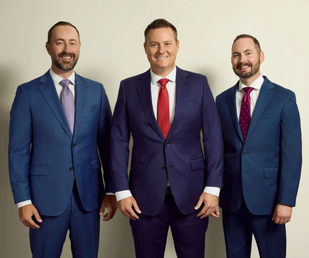 Three men in blue suits stand side by side against a plain light-colored background, all smiling and wearing ties.